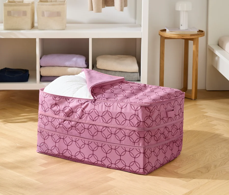 Ahşap zeminde, kısmen beyaz bir yorganla doldurulmuş, geometrik desenli pembe bir saklama çantası duruyor.