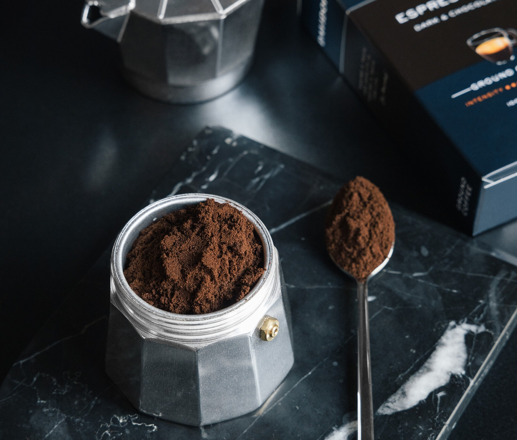 Mermer bir plaka üzerinde öğütülmüş kahveyle dolu gümüş bir espresso makinesinin, yanında öğütülmüş kahve bulunan bir kaşık ve bir paket DAVIDOFF CAFÉ Espresso 57 250g Gemahlen'in yakın çekimi.