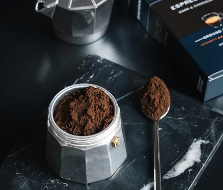 Mermer bir plaka üzerinde öğütülmüş kahveyle dolu gümüş bir espresso makinesinin, yanında öğütülmüş kahve bulunan bir kaşık ve bir paket DAVIDOFF CAFÉ Espresso 57 250g Gemahlen'in yakın çekimi.