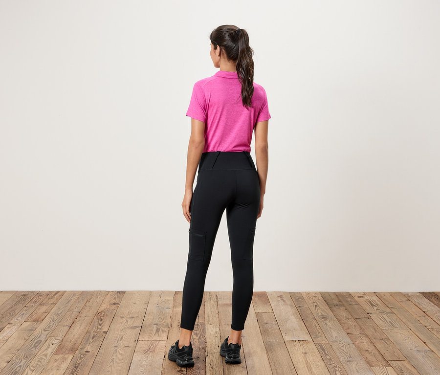 Bir kadın kameraya arkası dönük duruyor, pembe bir polo tişört ve cepli siyah tayt giyiyor.