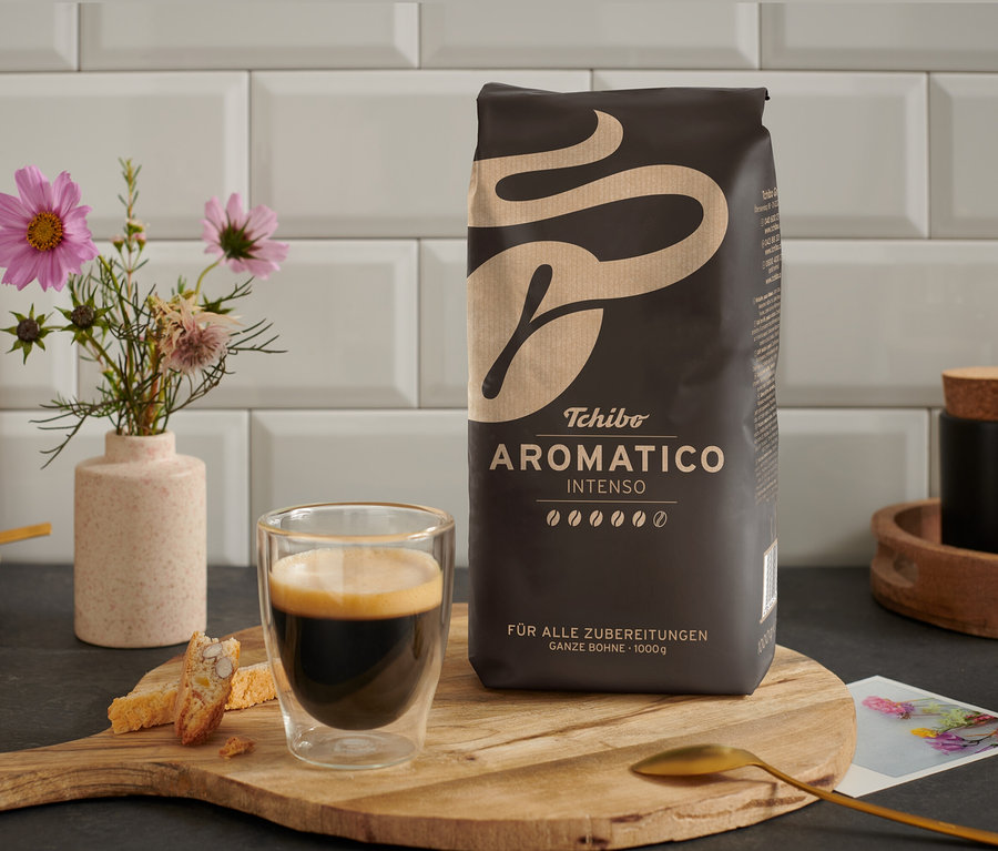 Ahşap bir masanın üzerinde bir fincan kahve, yanında bir paket Aromatico Intenso - 1 kg Çekirdek Kahve bulunmaktadır.