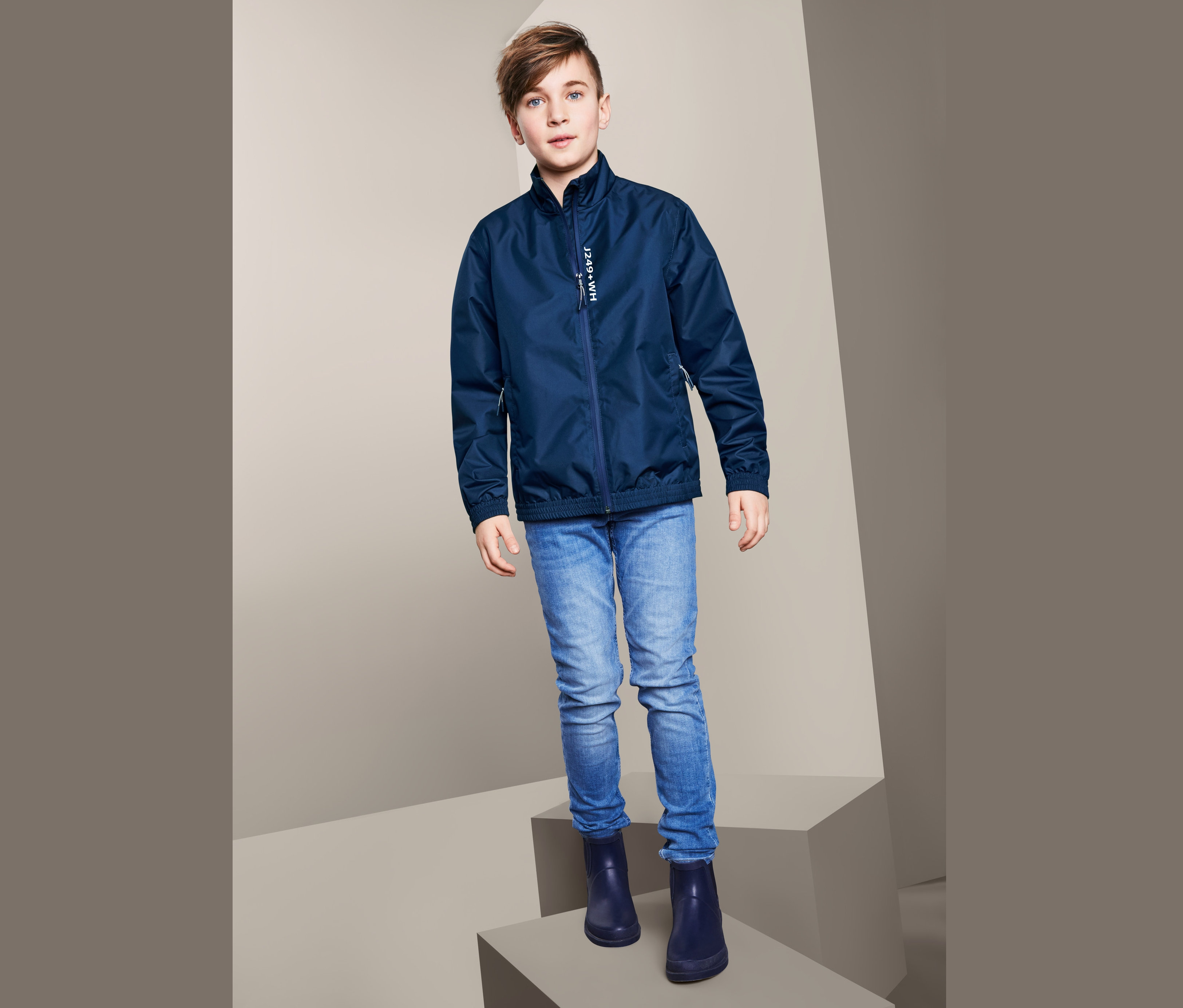 Bir erkek çocuk Bomber Ceket Tarzında Mevsimlik Ceket, mavi kot pantolon ve lastik çizme ile poz veriyor.