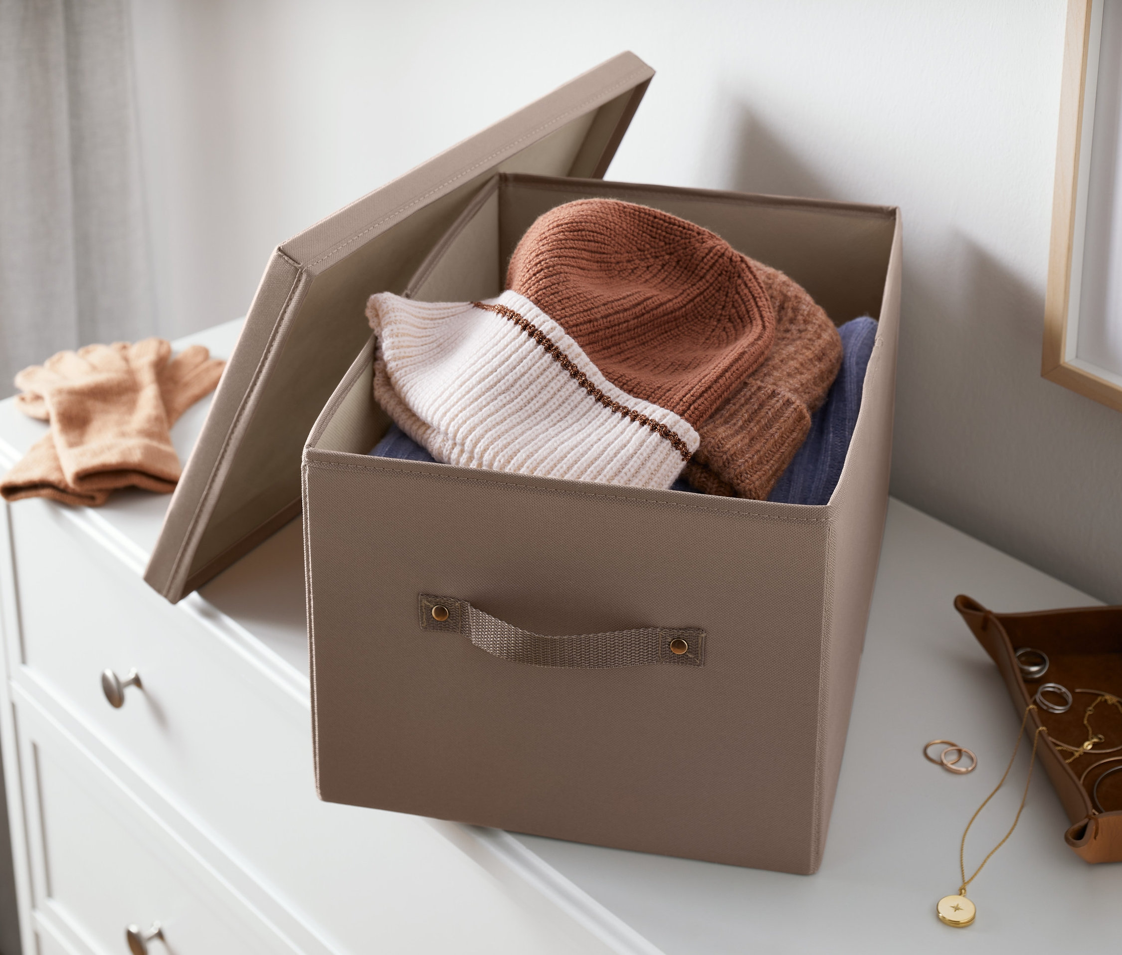 Şifonyerin üzerinde kapaklı, açık boz kahverengi düzenleme sandığı; içinde şapkalar ve giysiler var.