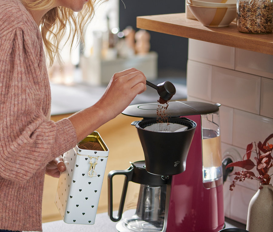 Sarışın saçlı bir kadın, Berry rengindeki »Let's Brew« Filtre Kahve Makinesine kahve döküyor.