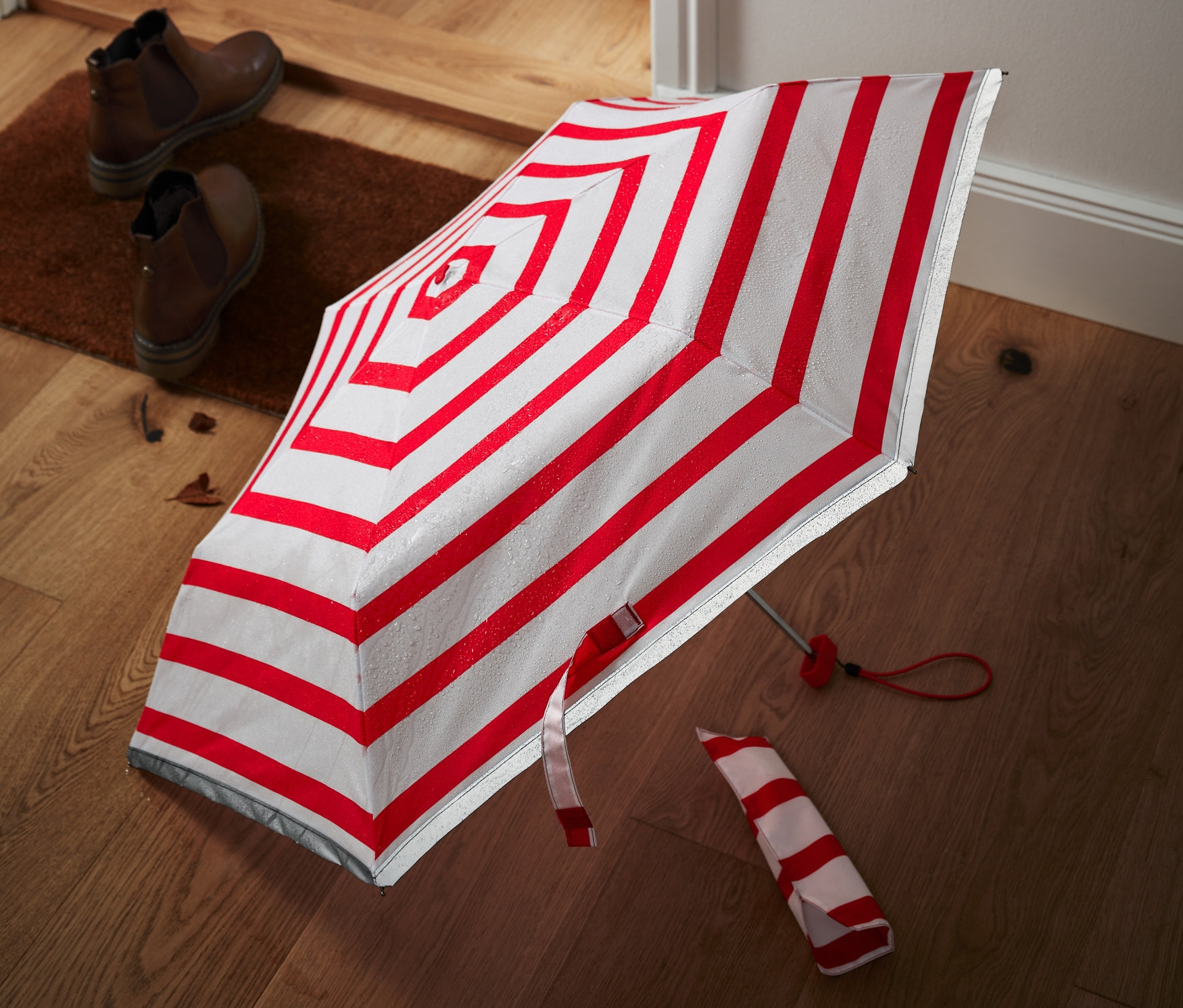 Kırmızı beyaz çizgili cep şemsiyesi, girişin yanındaki ahşap zeminde duruyor.