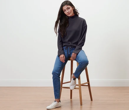 Bir kadın taburede oturuyor ve yarım balıkçı yaka sweatshirt, kot pantolon ve spor ayakkabı giyiyor.