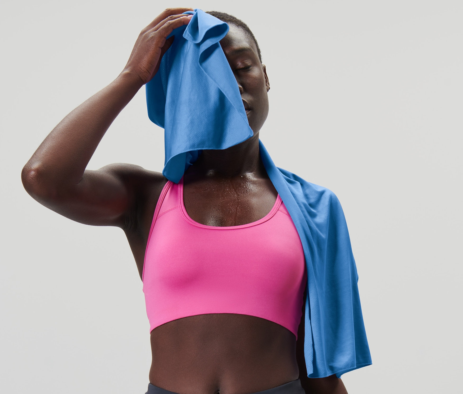 Kadın, gök mavisi rengindeki serinletici spor havlusuyla yüzünü siliyor. Pembe sporcu sütyeni ve koyu gri tayt giyiyor.