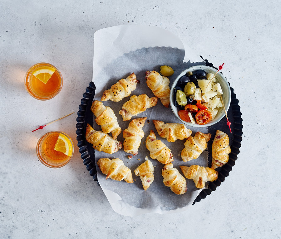 Kruvasanlar ve fırın tepsisinde zeytin ve domates içeren bir kase. Arka planda portakal dilimleri bulunan iki içecek.