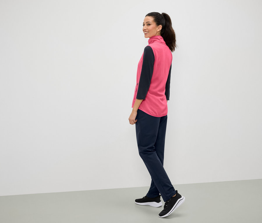 Bir kadın omzunun üzerinden gülümsüyor, pembe bir yelek, lacivert pantolon ve siyah spor ayakkabısı giyiyor.