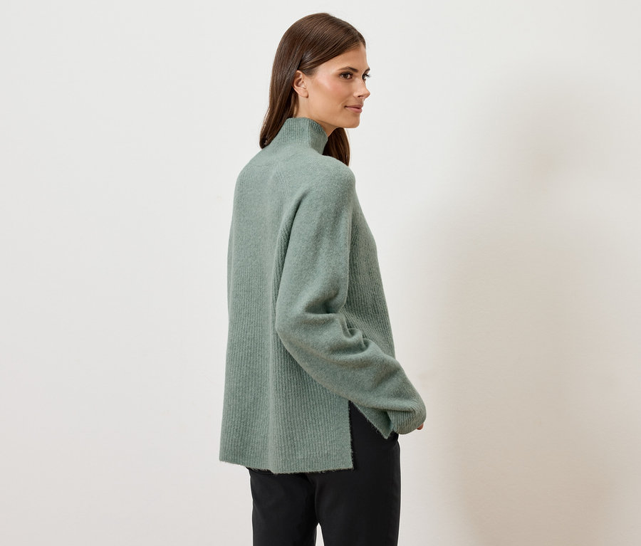 Kadın omzunun üzerinden bakıyor, yeşil balıkçı yaka kazak ve siyah pantolon giyiyor.