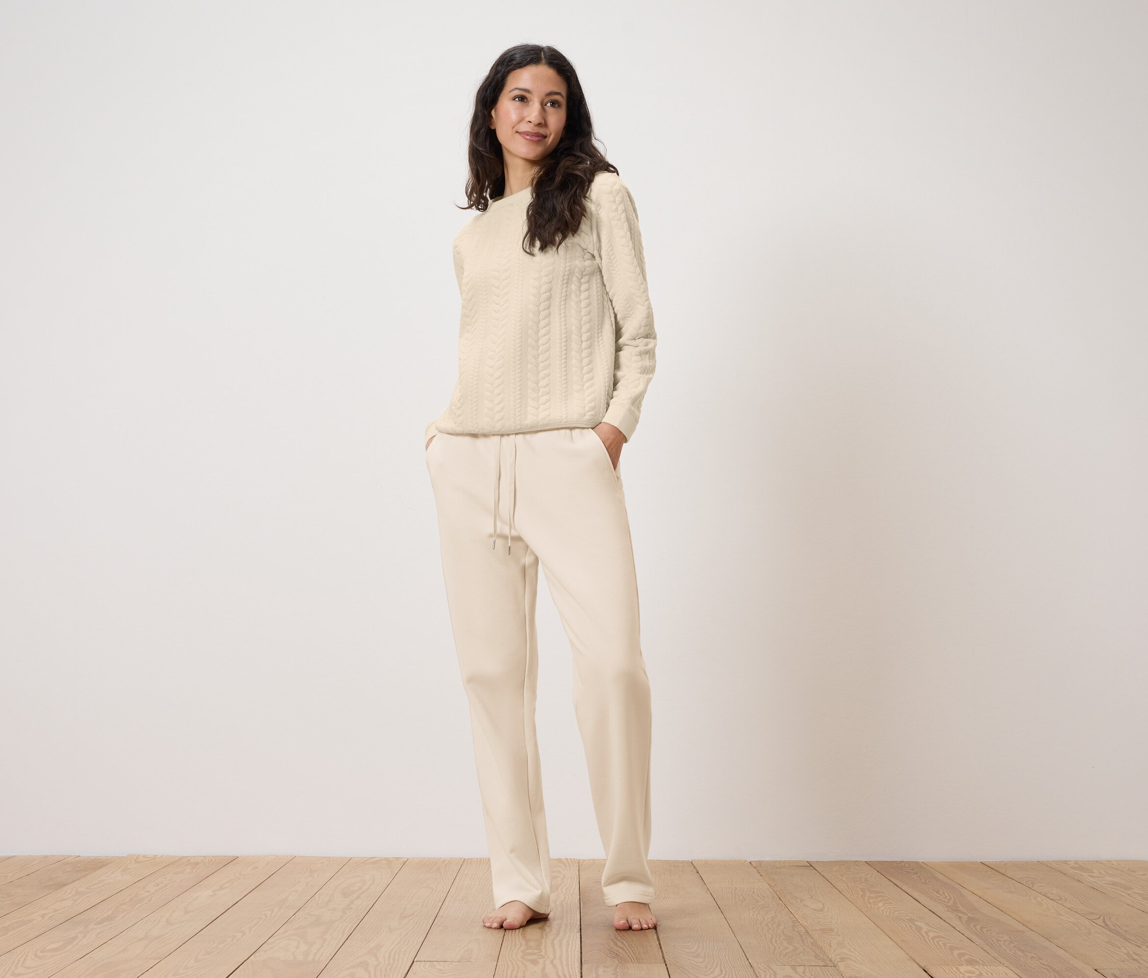 Bir kadın krem rengi eşofman altının ceplerinde elleri ve krem rengi örgü desenli bir kazakla ayakta duruyor.