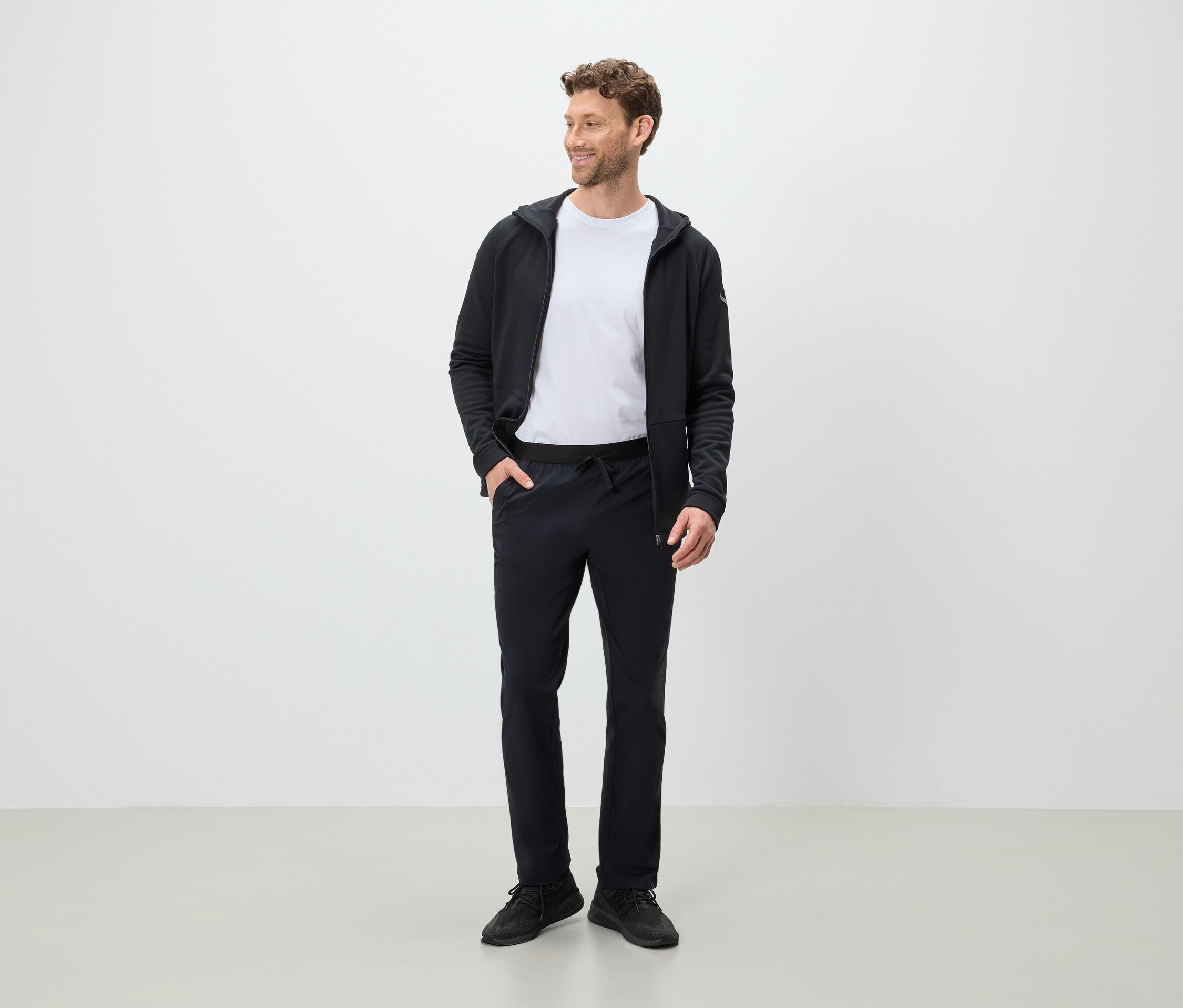 Bir adam ayakta duruyor ve yana bakıyor, üzerinde siyah pantolon, beyaz tişört ve siyah kapüşonlu bir ceket var.