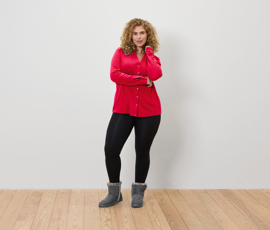 Kıvırcık saçlı bir kadın kırmızı bluz, siyah tayt ve gri botlarla beyaz bir duvarın önünde poz veriyor.