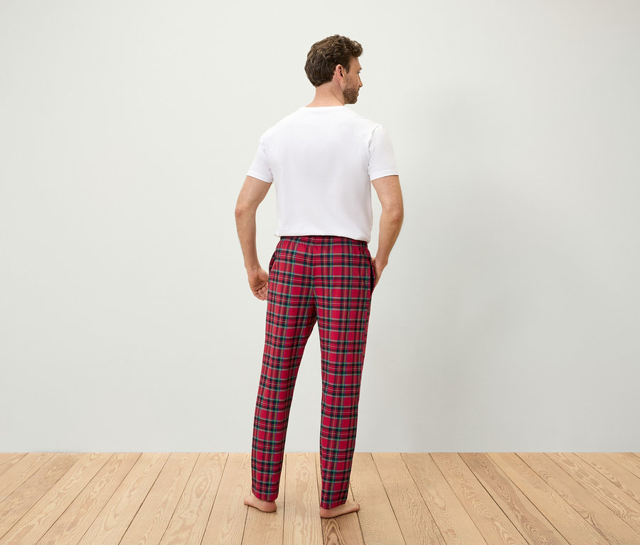Sırtı kameraya dönük bir adam, elleri kırmızı ekose pijama pantolonunun cebinde ve beyaz bir tişört giyiyor.
