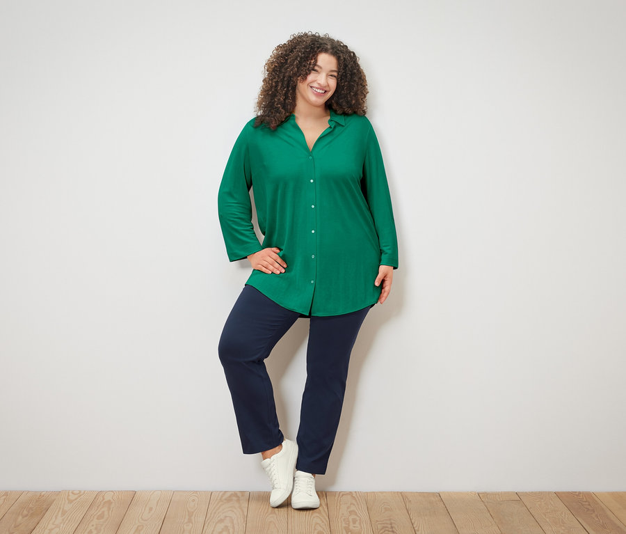 Kıvırcık saçlı, yeşil bluzlu, mavi pantolonlu ve beyaz spor ayakkabılı bir kadın beyaz bir duvarın önünde poz veriyor.