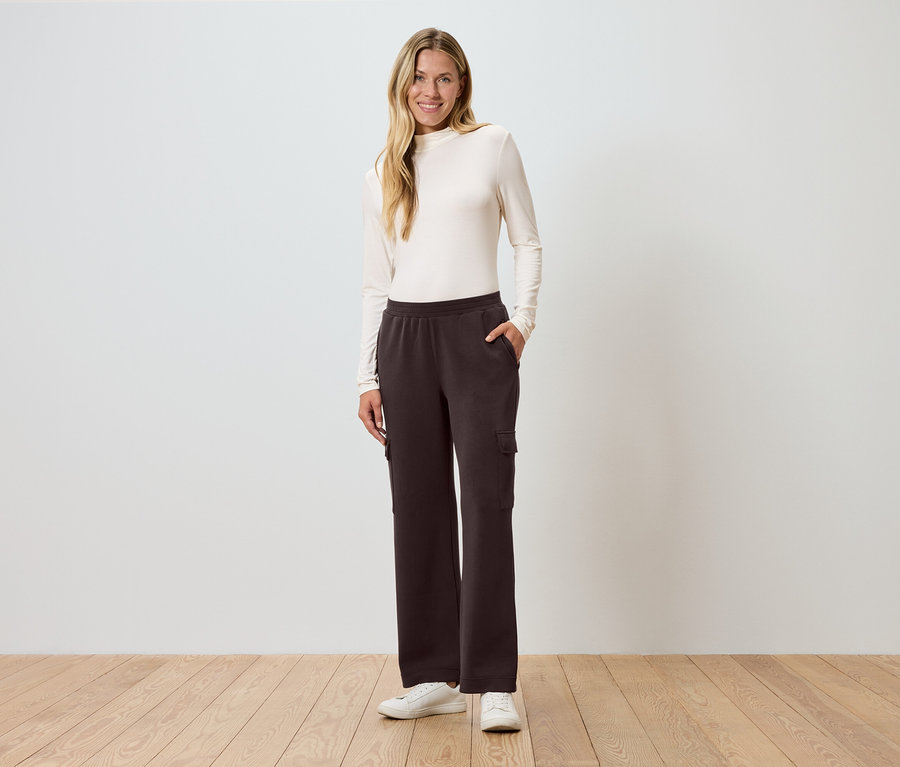 Kadın, eli cebinde bir duvarın önünde duruyor ve kahverengi kargo pantolon giyiyor.