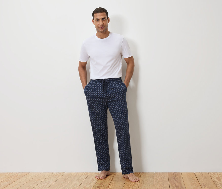 Bir adam beyaz bir duvarın önünde duruyor, beyaz bir tişört ve puantiyeli lacivert bir pijama altı giyiyor.