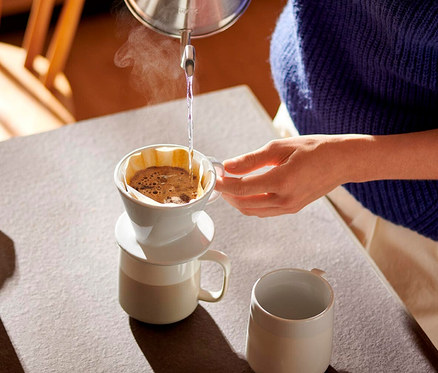 Bir kişi, bir kupanın üzerine yerleştirilmiş beyaz Filtre Kahve Aparatı, No. 101'e su ısıtıcısından sıcak su döküyor. Filtreden buhar yükseliyor. Masada başka bir kupa daha var.