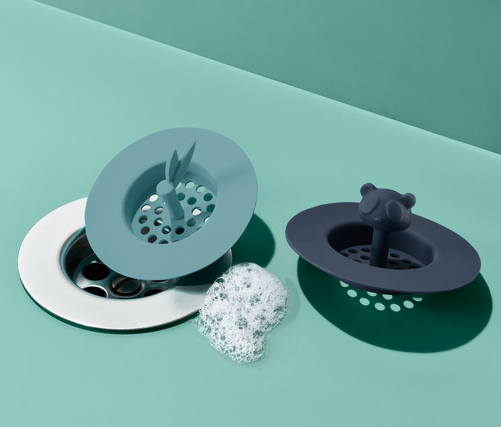 İki adet silikon lavabo süzgeci yeşil bir yüzeyde yan yana duruyor. Bir süzgeç açık mavi, diğeri panda desenli koyu mavi.