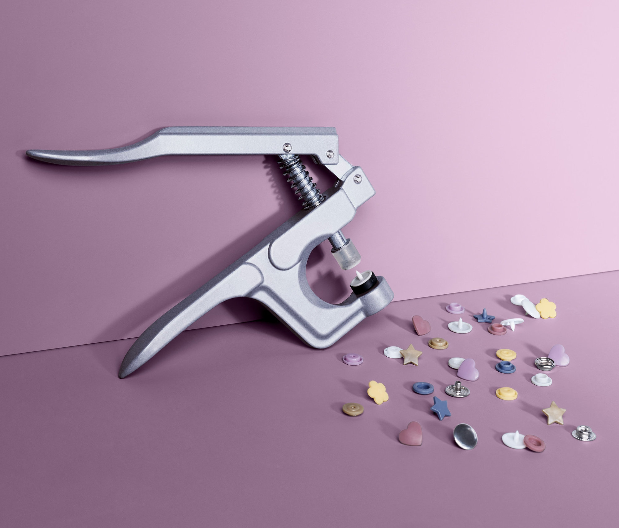 Lila bir zemin üzerinde gümüş bir pense ve farklı şekil ve renklerde bir çıt çıt düğme seti bulunmaktadır.