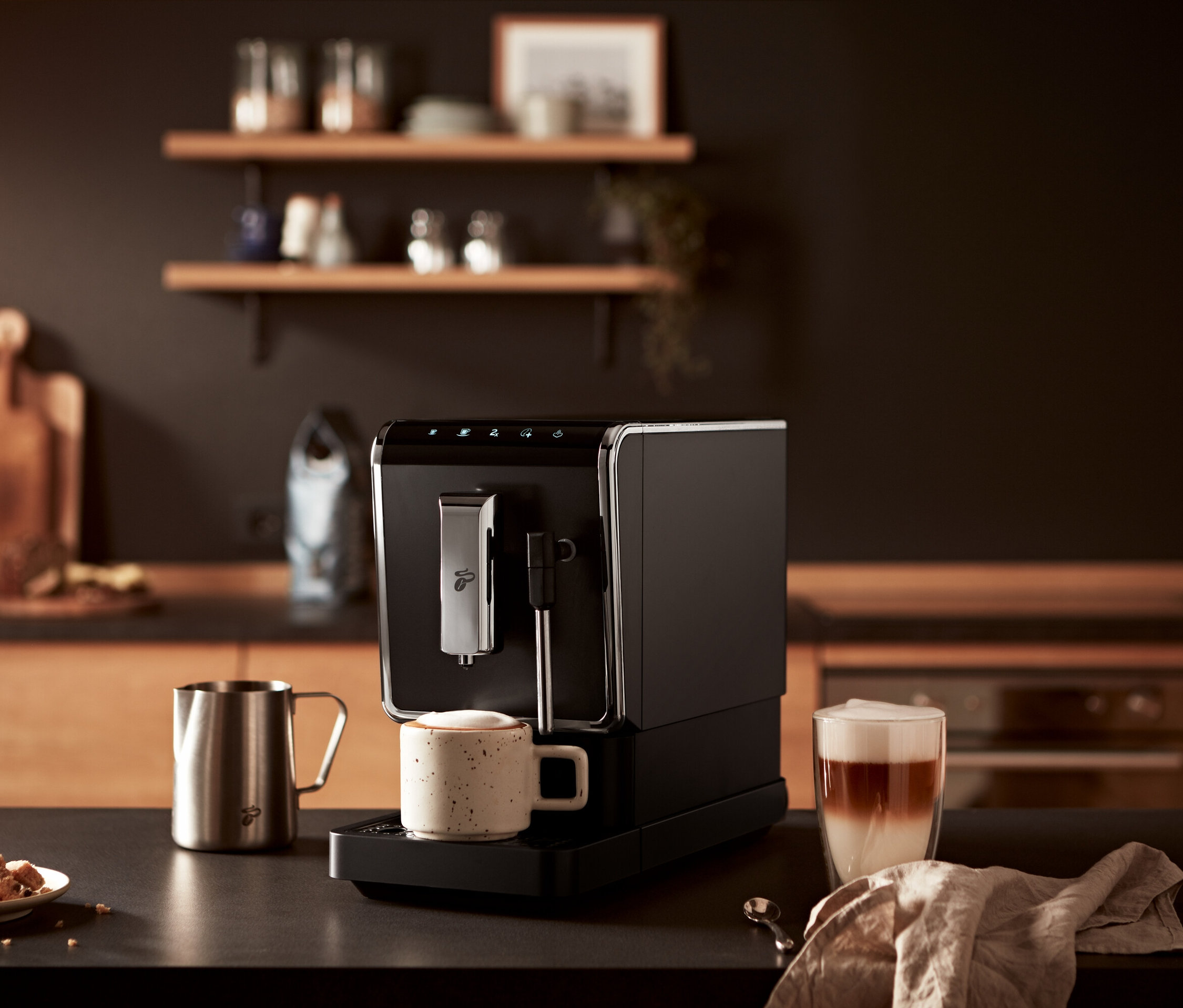 Bir masanın üzerinde antrasit rengi »Esperto Latte« Tchibo Tam Otomatik Kahve Makinesi, bir fincan kahve ve bir bardak latte bulunmaktadır.