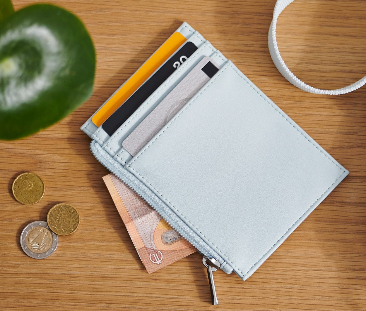 Ahşap bir masa üzerinde açık mavi bir cüzdan, madeni paralar ve bir çanta bulunmaktadır.