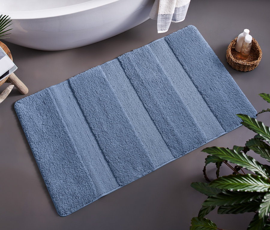 Mavi Wellness Banyo Paspası, yaklaşık 70 x 120 cm, bir bitkinin yanında yerde duruyor.