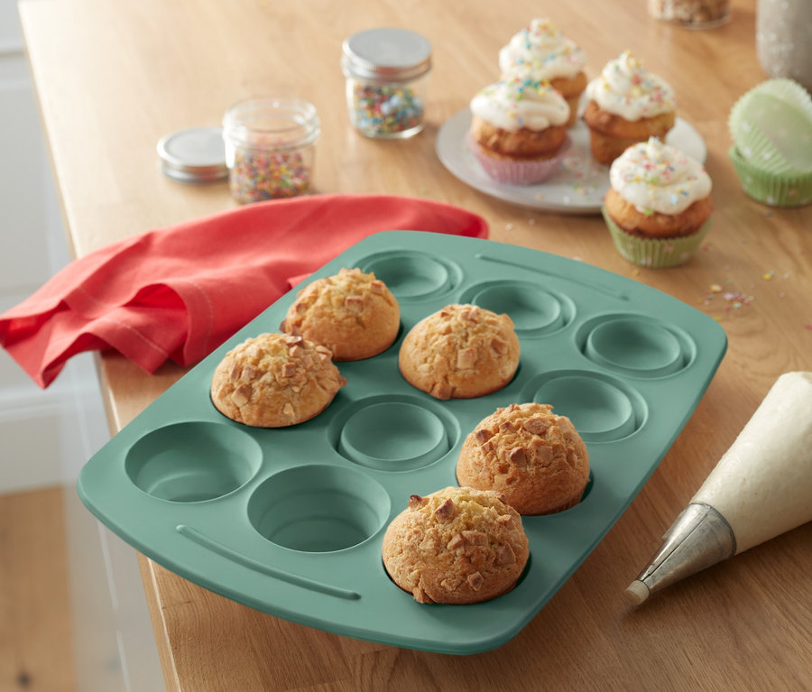 Ahşap bir masa üzerinde pişmiş muffinlerin bulunduğu silikon muffin kalıbı, krema torbası, renkli şekerlemelerin bulunduğu kavanozlar ve süslenmiş muffinlerin bulunduğu bir tabak bulunmaktadır.