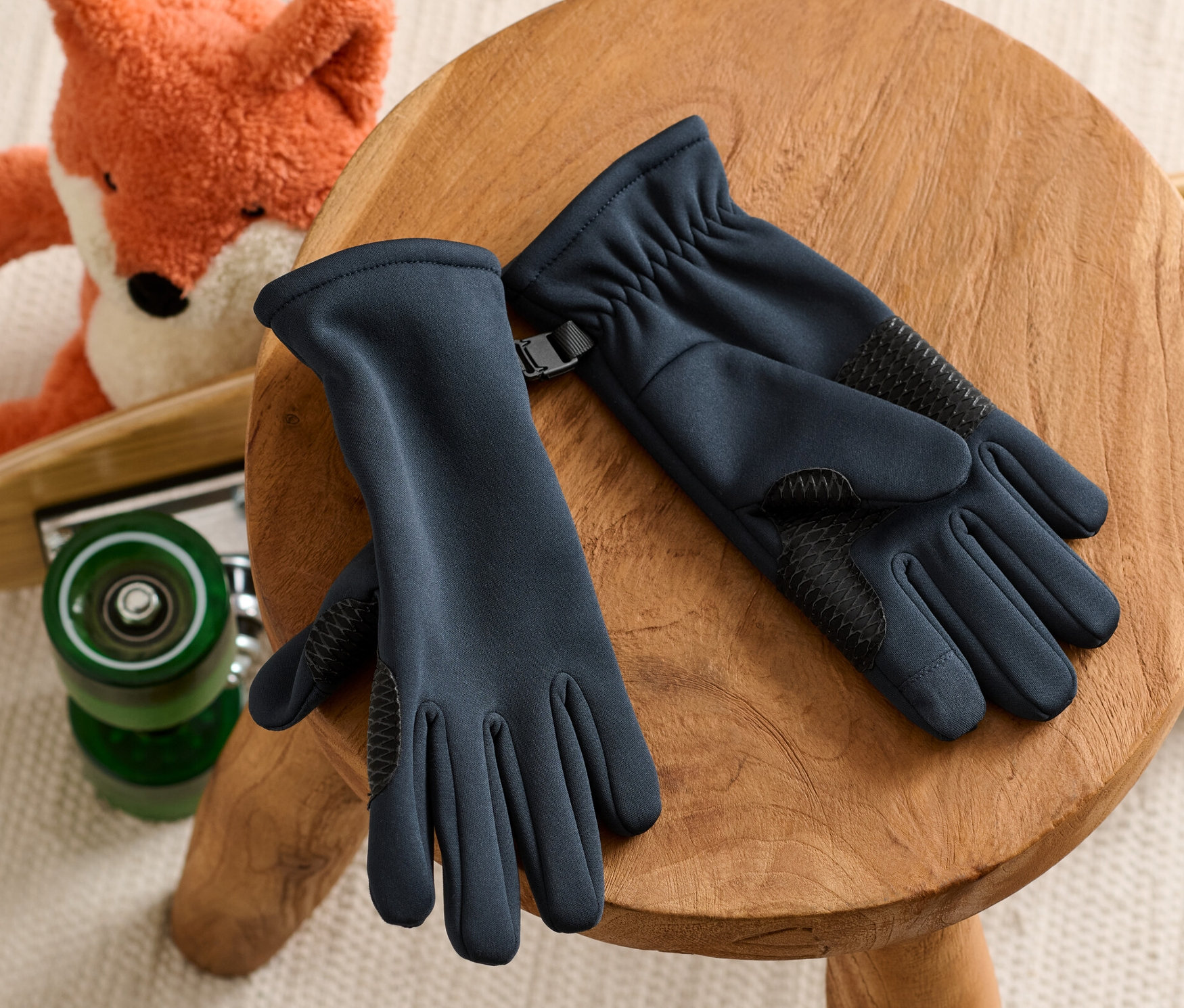 Çocuk softshell eldivenleri ahşap bir taburede duruyor, arka planda kaykay üzerinde peluş bir tilki var.