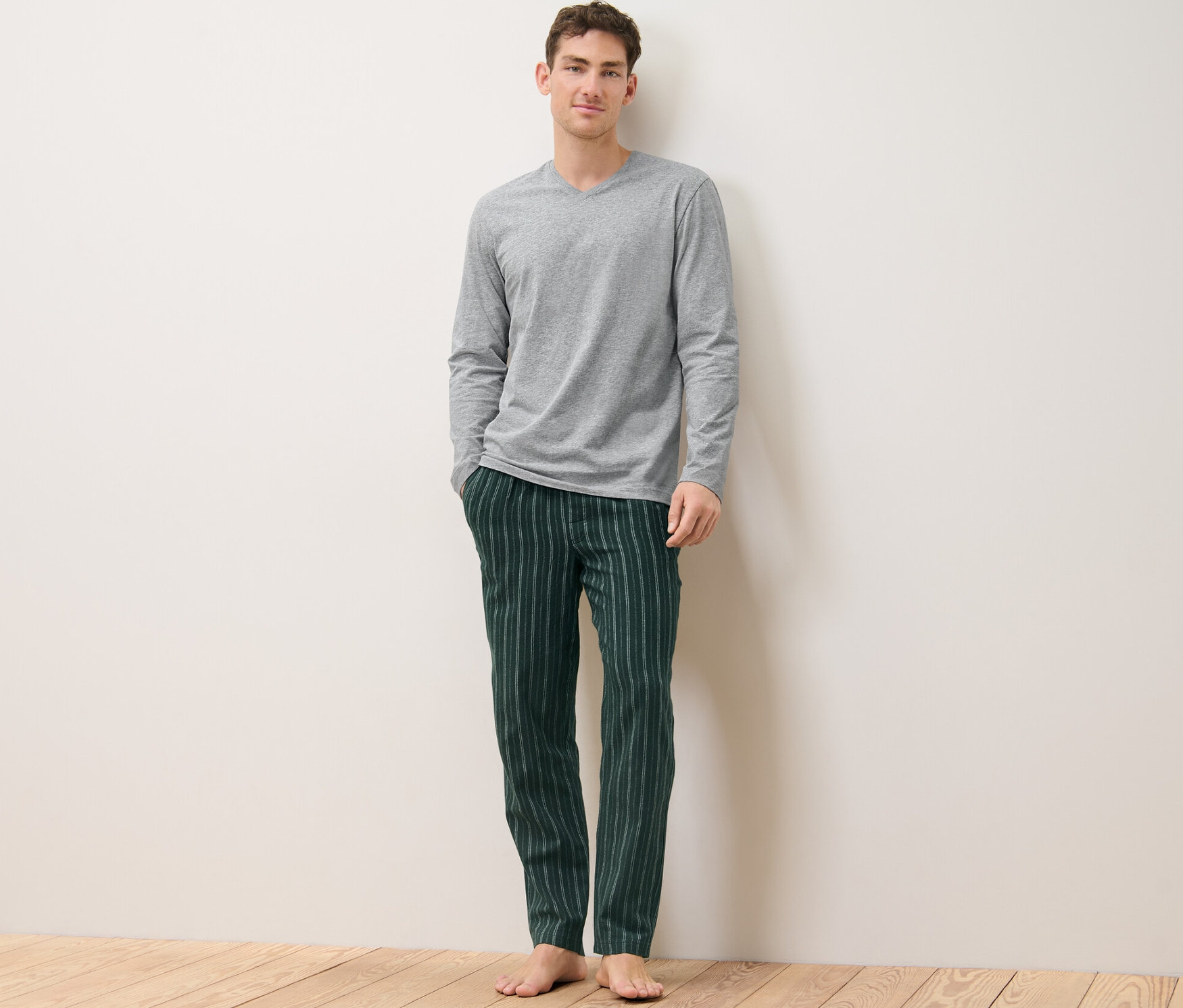 Adam ayakta duruyor, üzerinde gri, yeşil çizgili, pazen pijama takımı var.