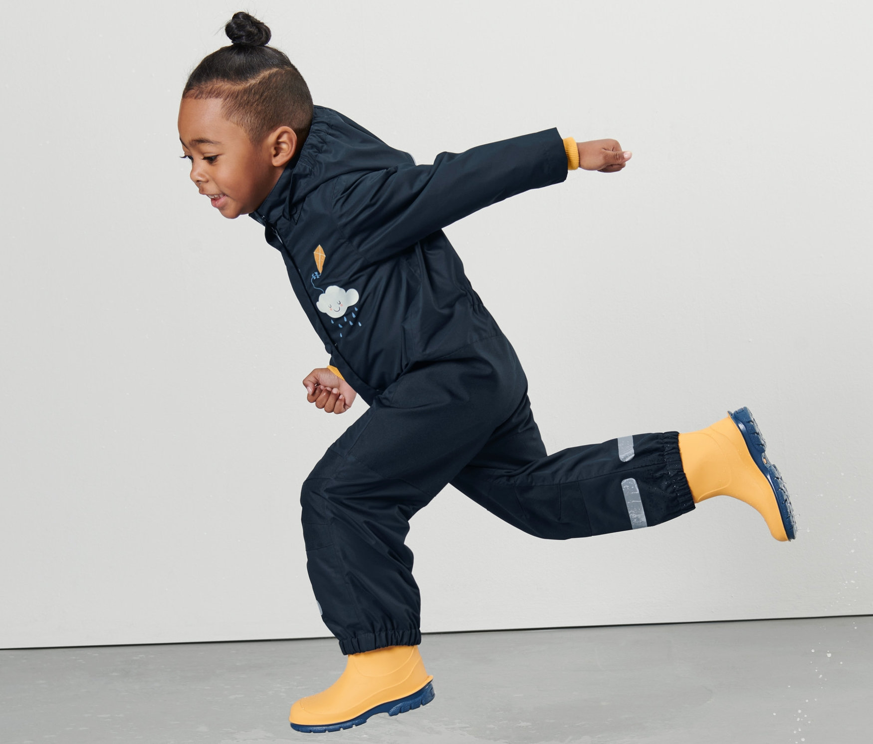 Küçük çocuk yağmur tulumu giymiş bir çocuk su birikintisinin içinde zıplıyor.