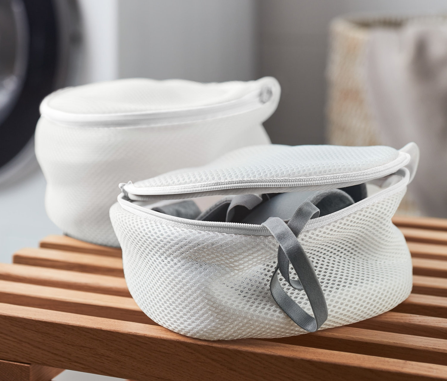 Ahşap bir bankta iki adet beyaz Sütyen Yıkama Filesi, arka planda bir çamaşır makinesi.