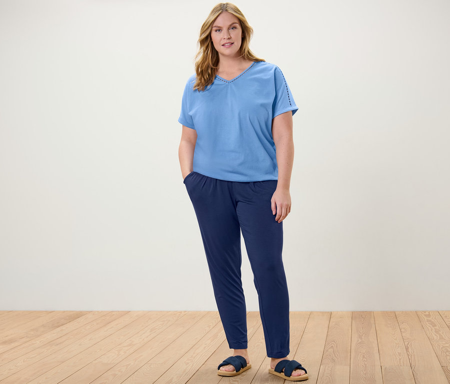 Bir kadın ayakta duruyor, açık mavi, kroşe şeritli bluz ve lacivert jarse pantolon giyiyor.