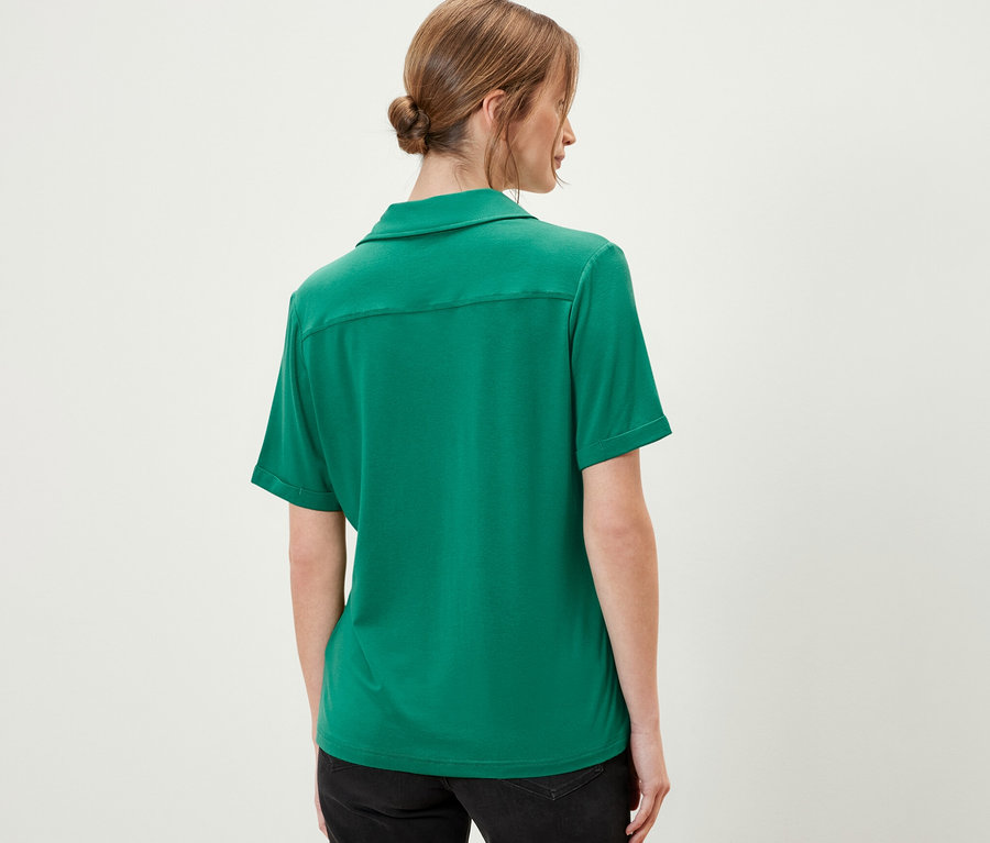 Arkadan zümrüt yeşili düğmeli bluz ve siyah şort giyen bir kadın görülüyor.