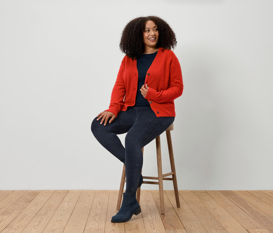 Bir kadın taburede oturuyor, kırmızı bir hırka, mavi kot pantolon ve mavi botlar giyiyor.