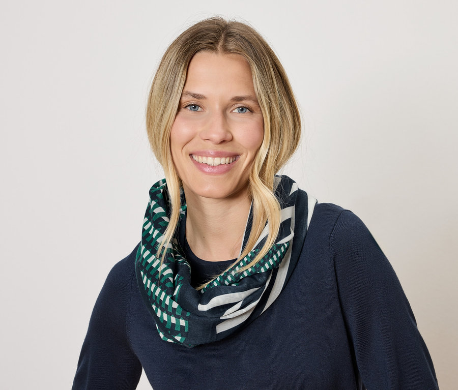 Sarışın saçlı, gülümseyen bir kadın lacivert bir kazak ve yeşil ve beyaz geometrik desenli bir eşarp takıyor.