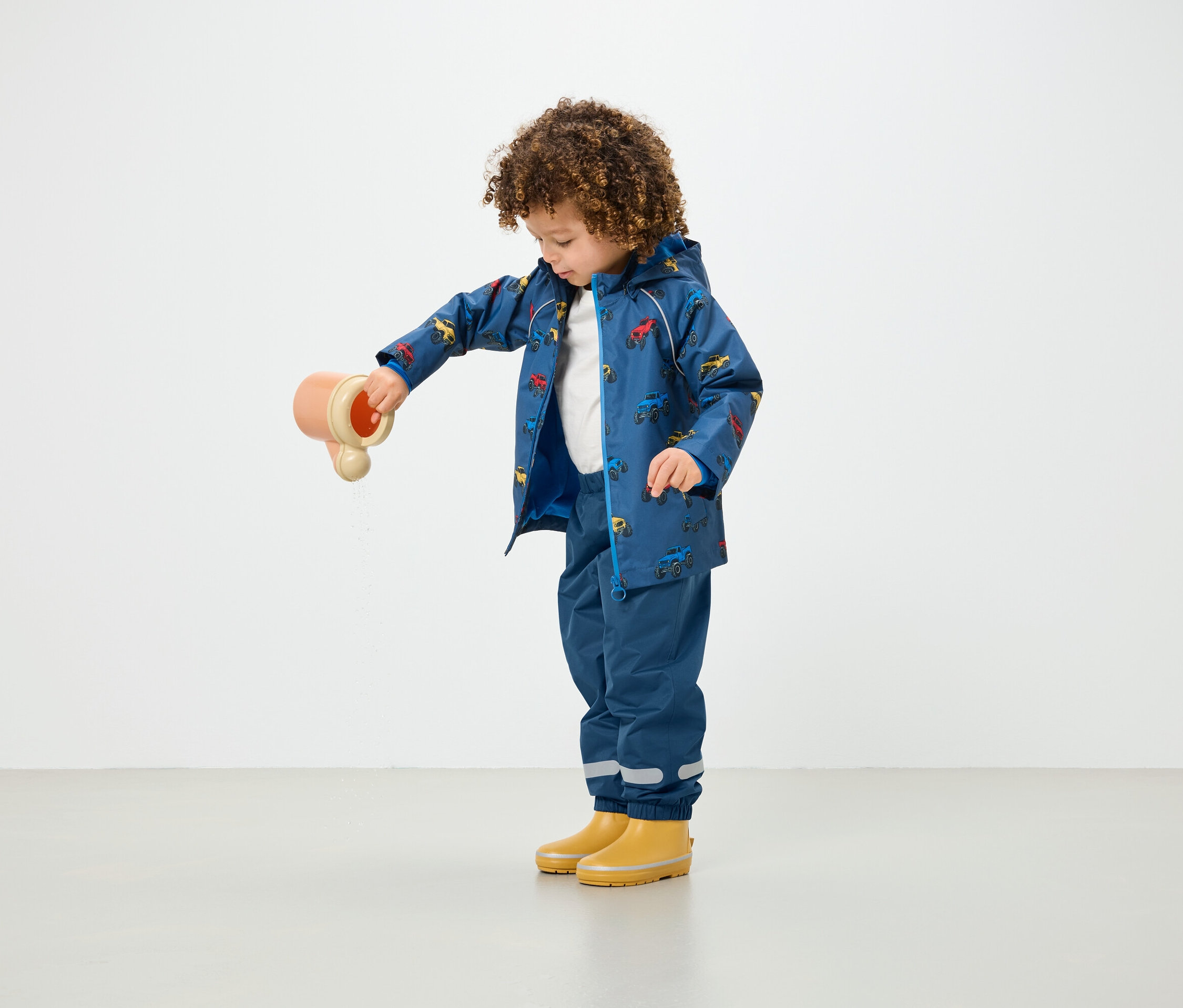 Kıvırcık saçlı bir çocuk, baskılı mavi bir yağmurluk ve mavi pantolon giyerken bir oyuncaktan su döküyor.