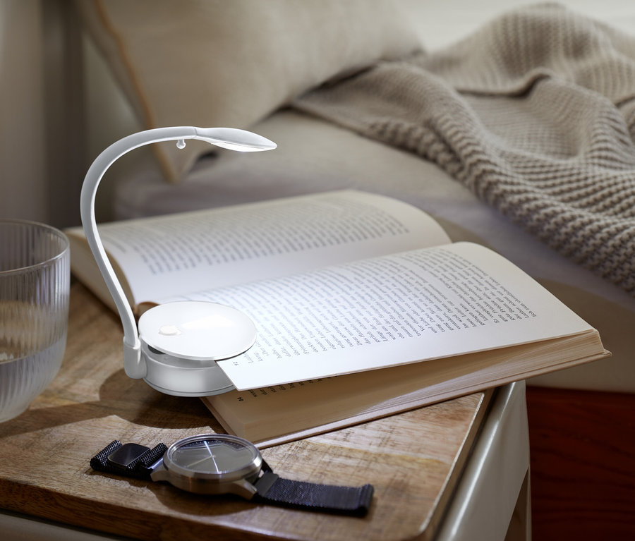 Bir komodinin üzerinde küçük bir lamba ile aydınlatılmış açık bir kitap, yanında bir bardak su ve bir saat duruyor.