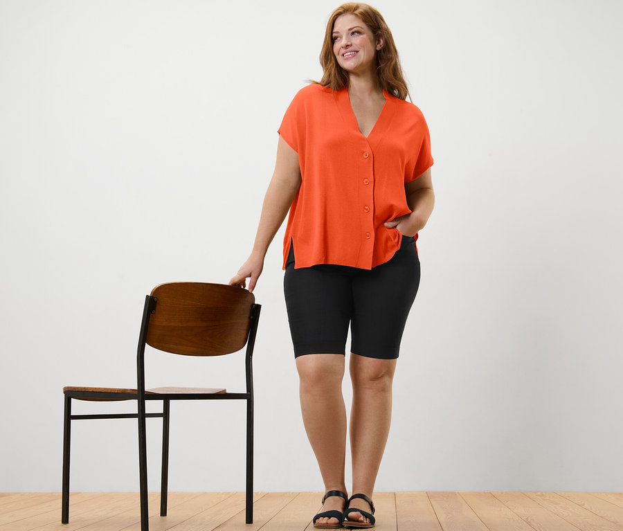 Bir kadın sandalyenin yanında duruyor ve turuncu kısa kollu bir hırka ve siyah Bengal kısa şort giyiyor.