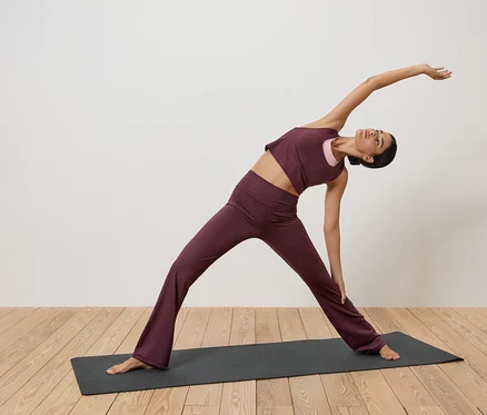 Bordo renkli spor kıyafetleri giyen bir kadın, beyaz duvar önünde ahşap zemin üzerinde yoga matında esniyor.