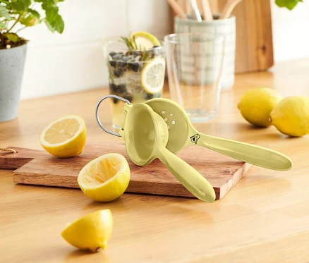Ahşap bir masanın üzerinde birkaç limon, limon sıkacağı ve bir bardak limonata duruyor.