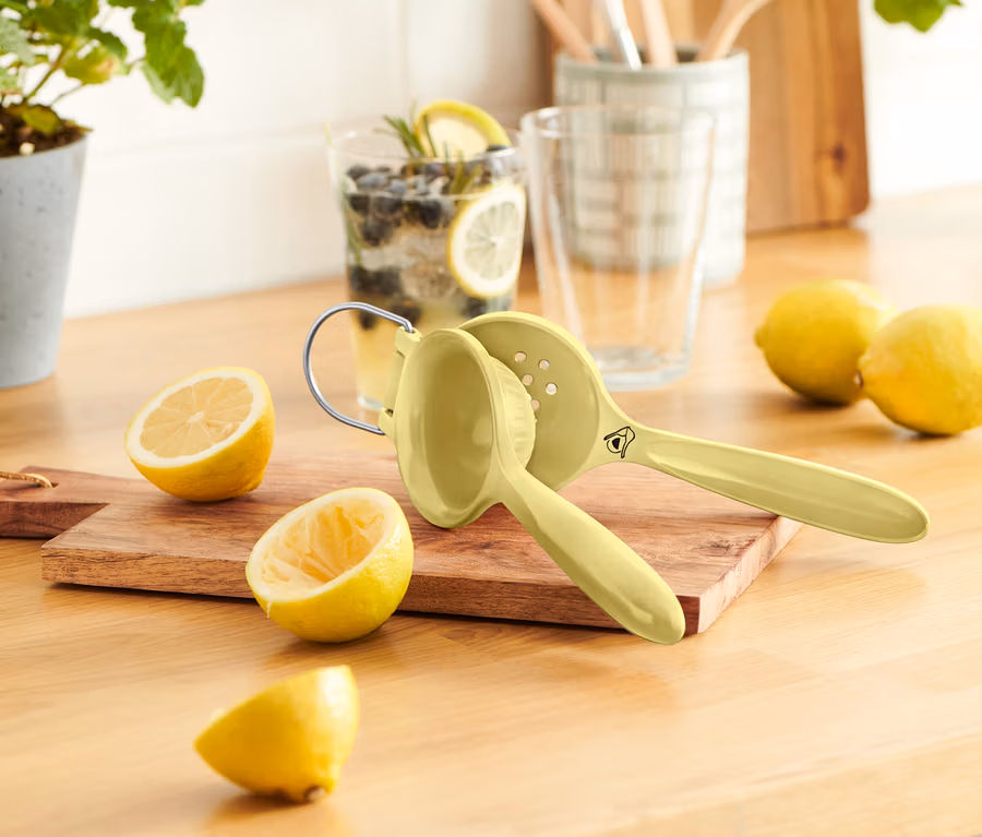 Ahşap bir masanın üzerinde birkaç limon, limon sıkacağı ve bir bardak limonata duruyor.