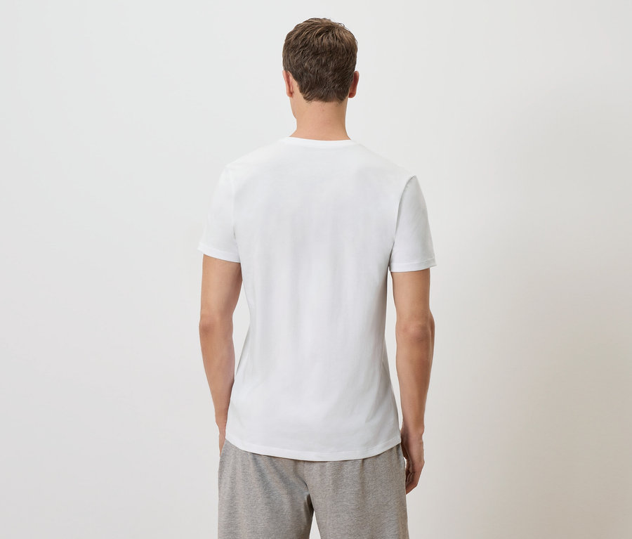 Bir adam kameraya arkası dönük duruyor, beyaz bir tişört ve gri şort giyiyor.