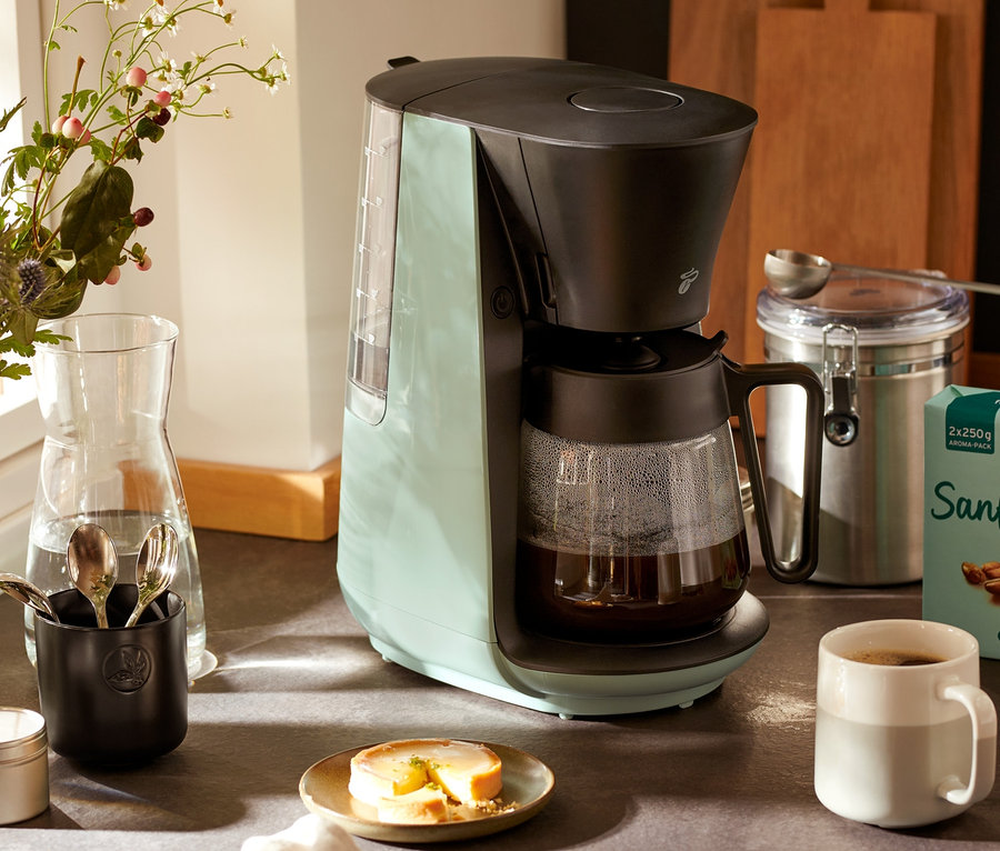Açık yeşil »Let's Brew« Filtre Kahve Makinesi, bir fincan kahve ve bir çiçek vazosunun yanında bir mutfak tezgahında duruyor.