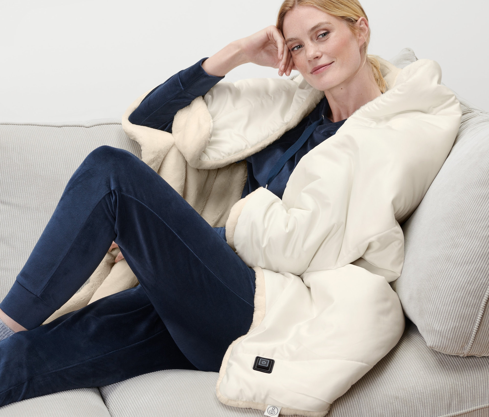 Kadın kanepede oturuyor, üzerinde şarjlı ısıtıcı panço var.