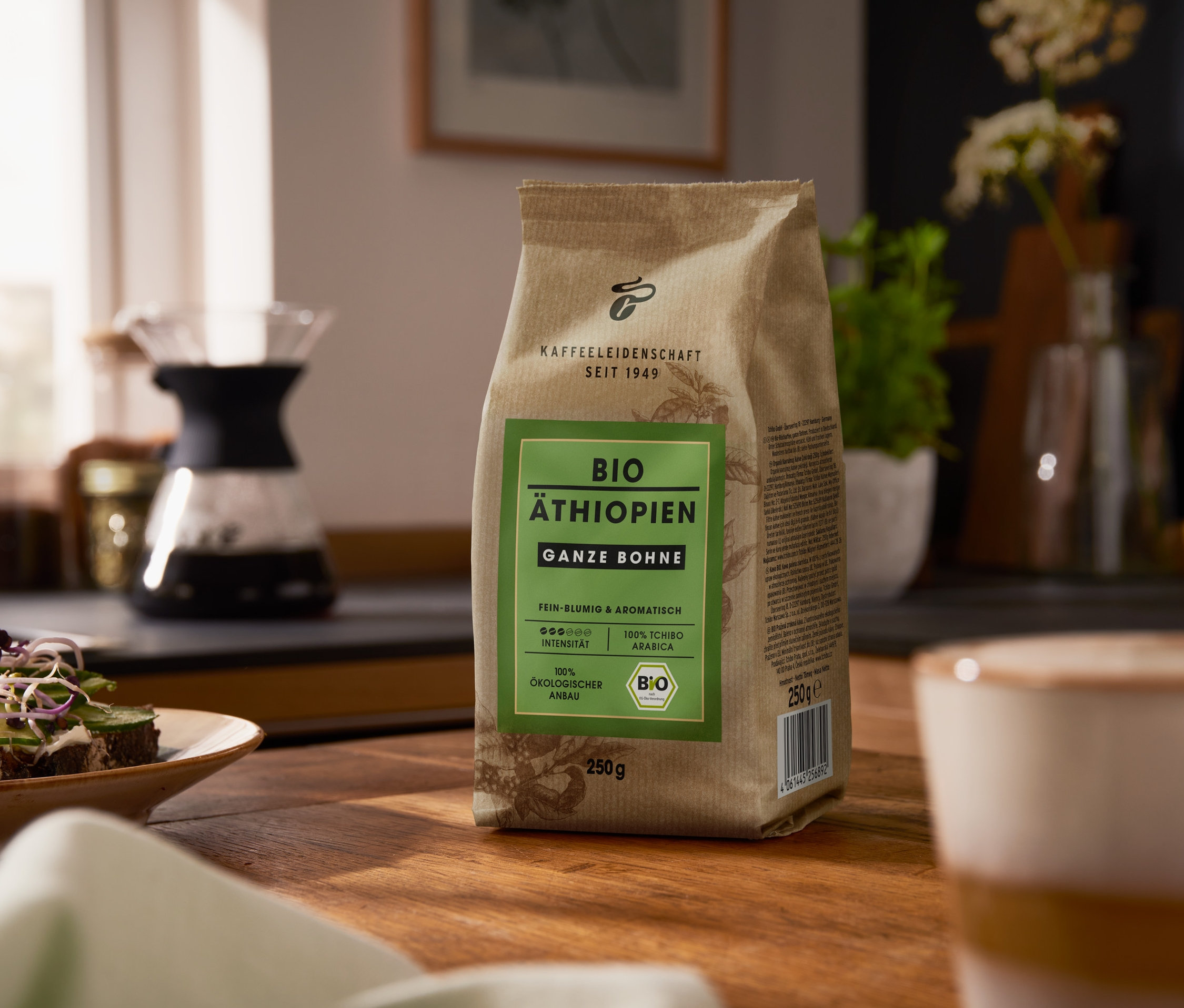 Ahşap bir masanın üzerinde bir paket Bio Äthiopien - 250 g Ganze Bohne duruyor. Arka planda bir kahve makinesi var.