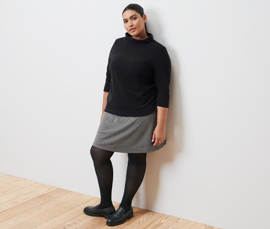 Duvara yaslanmış bir kadın, siyah 3/4 kollu sweatshirt, sweat etek, siyah külotlu çorap ve deri loafer giyiyor.