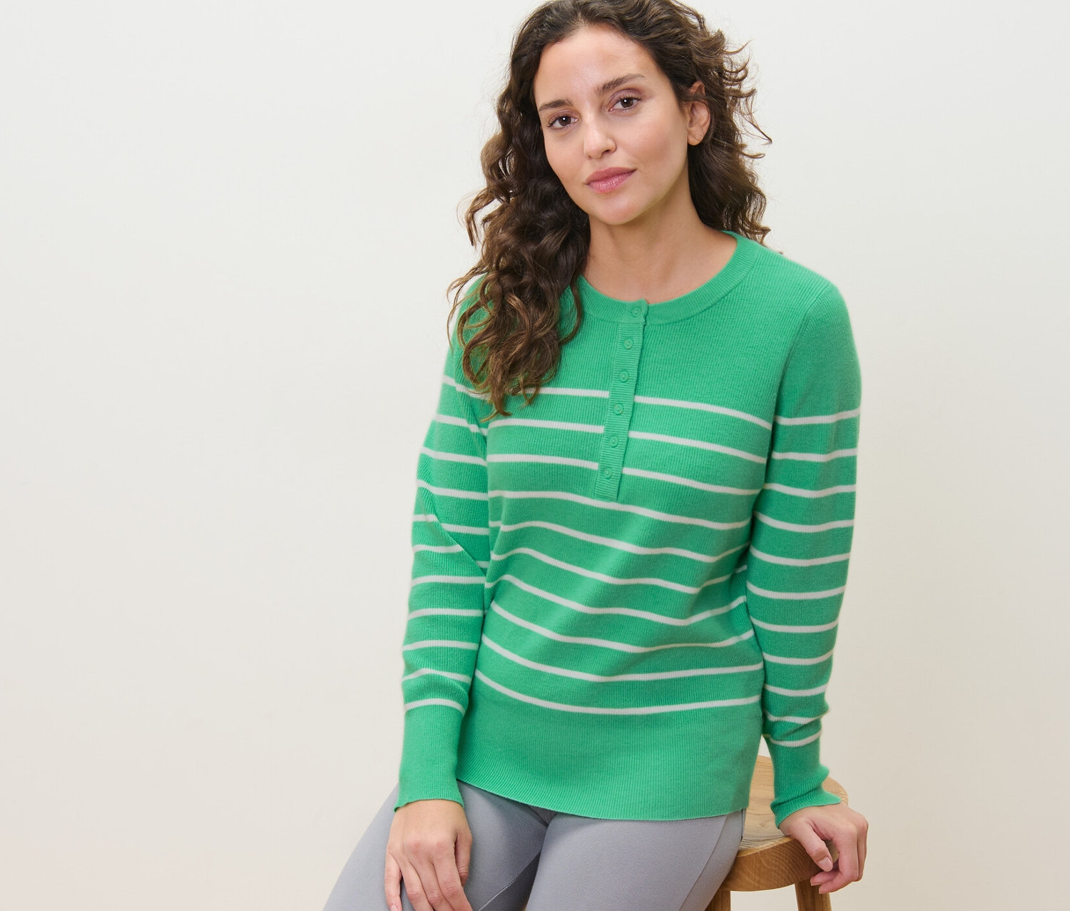 Bir kadın taburede oturuyor ve çizgili, ince örgülü bir kazak giyiyor.