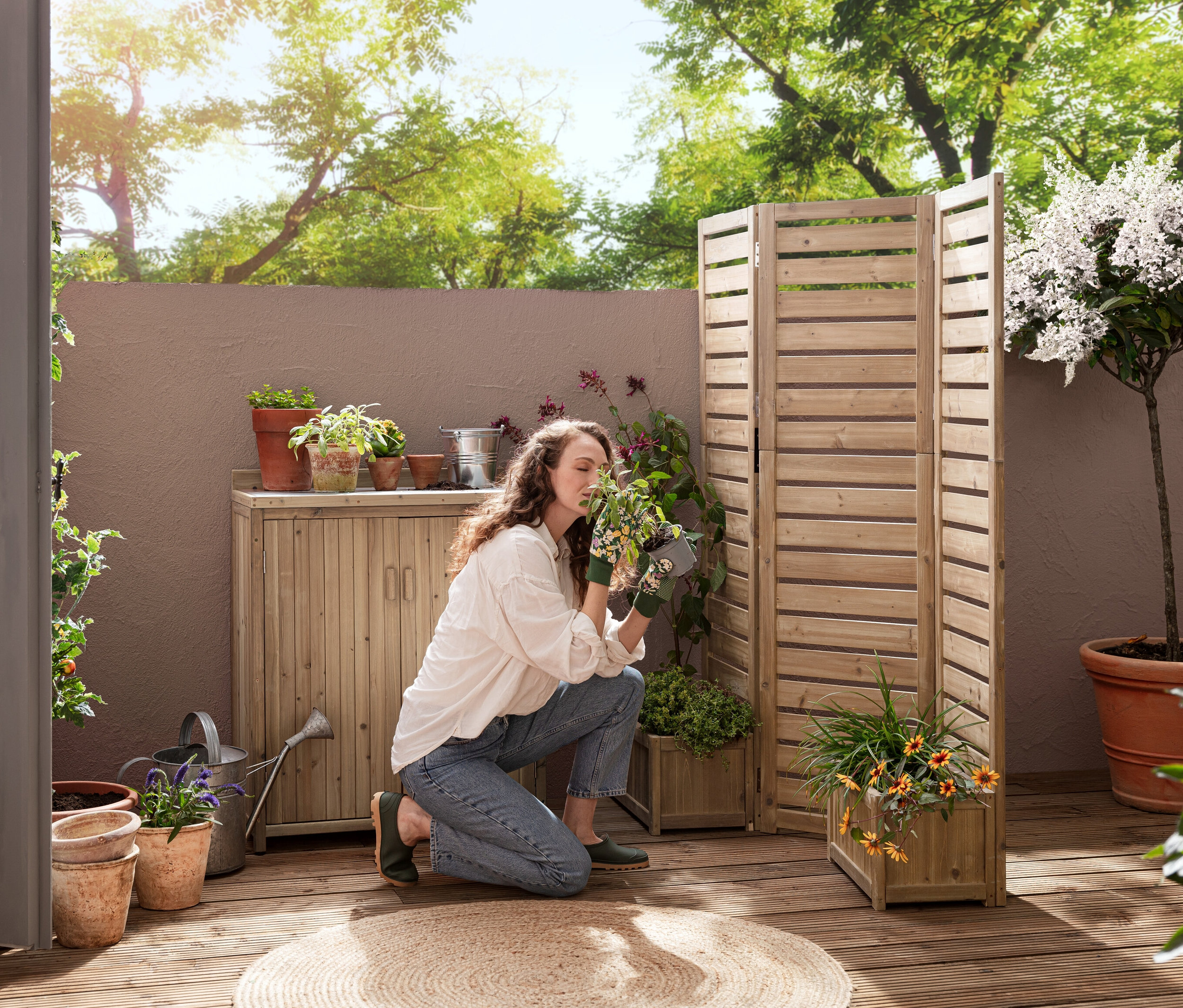 Bir kadın terasta diz çökmüş, Saksı Kutulu Esnek Paravan »Tinus« ve Küçük Bahçe Dolabı »Tinus« yakınında bir bitkiyi kokluyor.