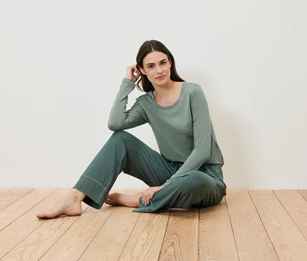 Kadın ahşap zeminde oturuyor ve yeşil bir pijama giyiyor.